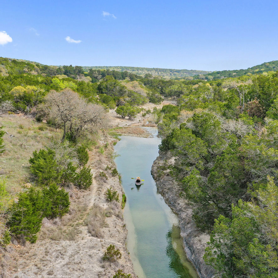 An elevated view of a woman kayaking in a small, serene stream surrounded by trees in the Texas Hill Country.