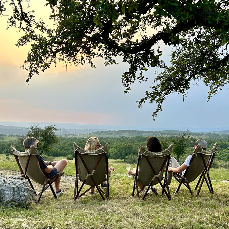 Four people sit in folding chairs on a grassy hill, facing a scenic view of rolling green hills and a pastel-colored sky at sunset. The branches of a nearby tree frame the top of the image, adding to the peaceful, outdoor setting as the group enjoys the tranquil landscape.