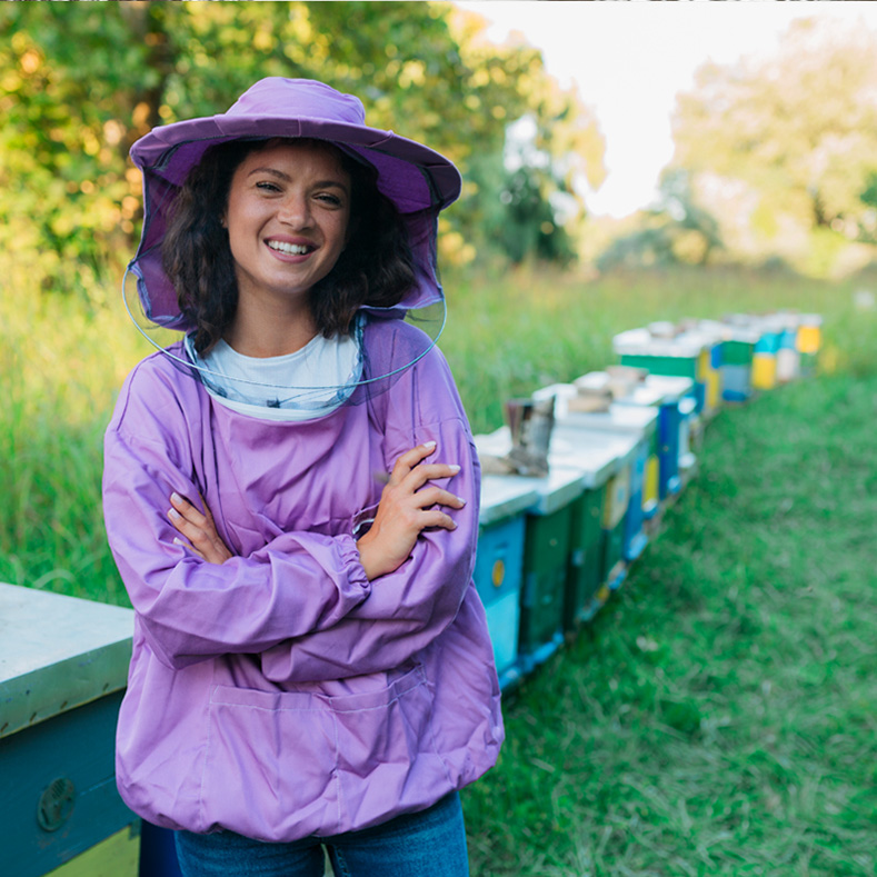 A smiling woman wearing a purple beekeeping suit and hat with a protective mesh stands with her arms crossed in front of a row of colorful beehives in a grassy field. The background shows a bright, natural setting with trees and greenery, suggesting she is tending to the beehives in an outdoor environment.