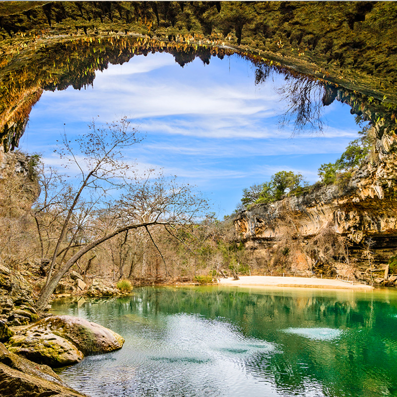 A serene natural pool surrounded by tall, rocky cliffs, with a large overhanging cave-like formation framing the top of the image. The clear water reflects the trees and sky, while bare tree branches lean over the water's edge. The scene is peaceful, with the blue sky and scattered clouds visible through the opening in the rocky arch, suggesting a hidden oasis in a remote landscape.