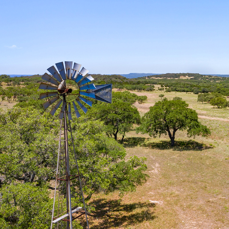 A metal windmill stands tall in the foreground, surrounded by lush green trees and open grassy fields under a clear blue sky. In the distance, rolling hills and patches of trees stretch toward the horizon, creating a peaceful rural landscape.