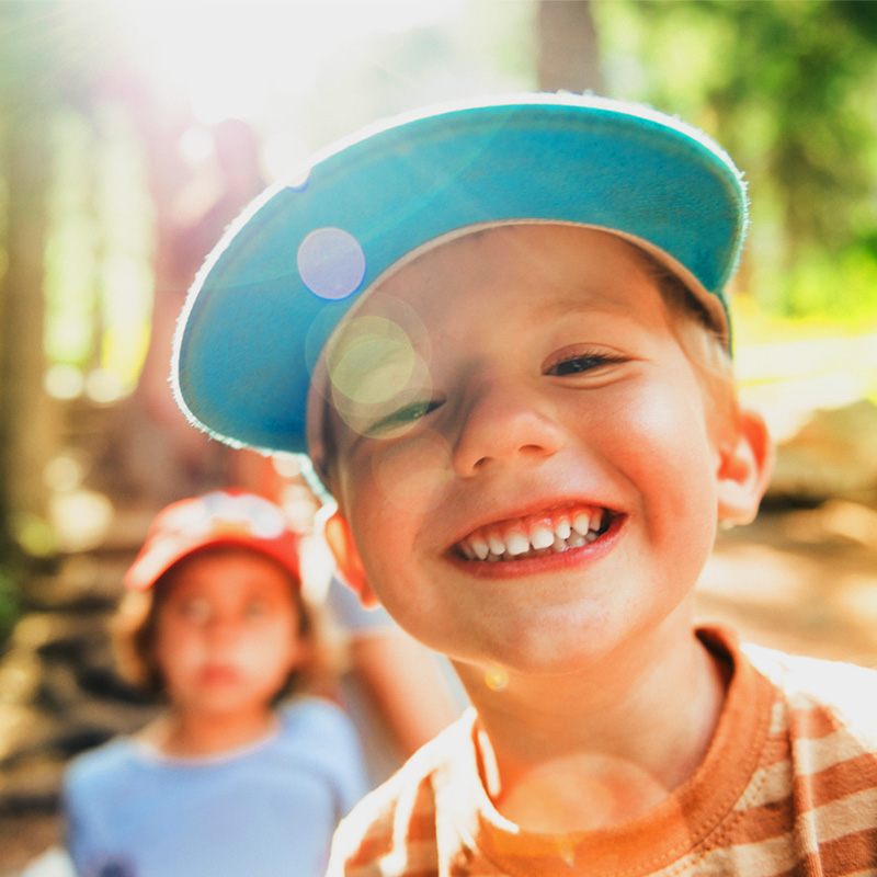 A young child smiling brightly while wearing a blue cap, with sunlight creating lens flare effects in the background. The child is wearing an orange-striped shirt, and another child in a red cap and blue shirt is visible in soft focus behind them. The setting appears to be outdoors, possibly in a forest, with warm sunlight and greenery surrounding them.