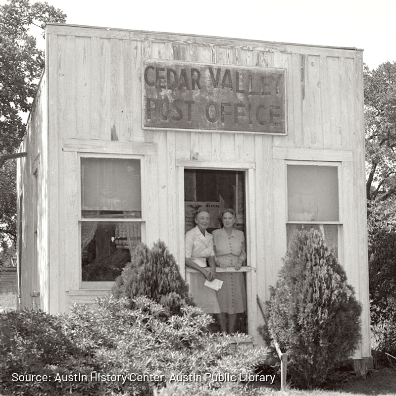 A historical photograph of two people standing at the entrance of the Cedar Valley Post Office. The small wooden building has a simple sign above the doorway that reads 'Cedar Valley Post Office.' The individuals are standing in front of the door, and some shrubs frame the building in the foreground. The image captures a quaint and intimate moment of a bygone era in a rural community.