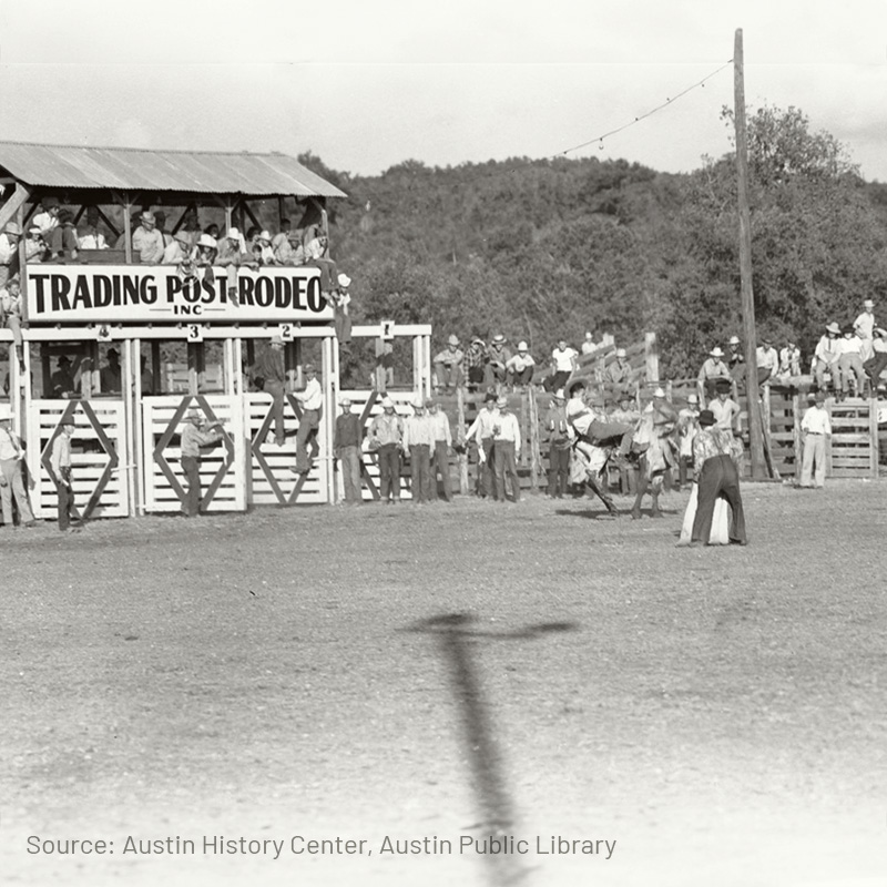 A vintage photograph of a rodeo scene, with a rider attempting to stay on a bucking horse in the center of the arena. Spectators are gathered in bleachers and a wooden stand labeled 'Trading Post Rodeo, Inc.' Cowboys and rodeo participants are seen nearby, watching the event unfold. The photograph captures the essence of a traditional rodeo atmosphere.