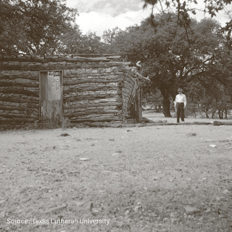 A sepia-toned photograph of a rustic log cabin with a single door, situated in a wide, open area. In the distance, a man wearing a hat and formal clothing stands near the cabin, with large trees providing a shaded backdrop. The scene reflects a historical and rural setting.