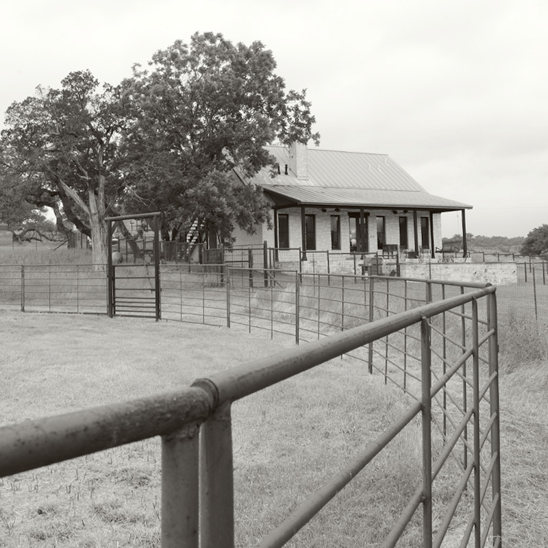 A black-and-white photograph of a small farmhouse surrounded by metal fencing. The house has a metal roof, a wide porch, and several windows, with a large tree beside it. The fencing encloses a grassy area, and the landscape beyond the house is open, suggesting a quiet rural setting.