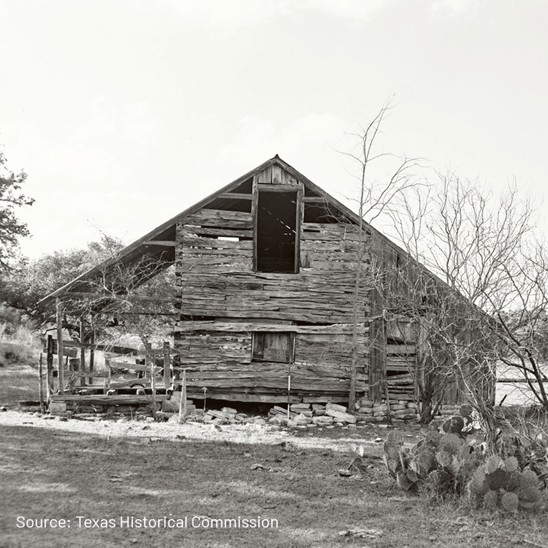 A black-and-white photograph of an old, weathered wooden structure with missing planks and a partially collapsed roof. The building appears abandoned and is surrounded by sparse trees and cacti. The rustic, historical nature of the structure suggests it may be an old barn or cabin.