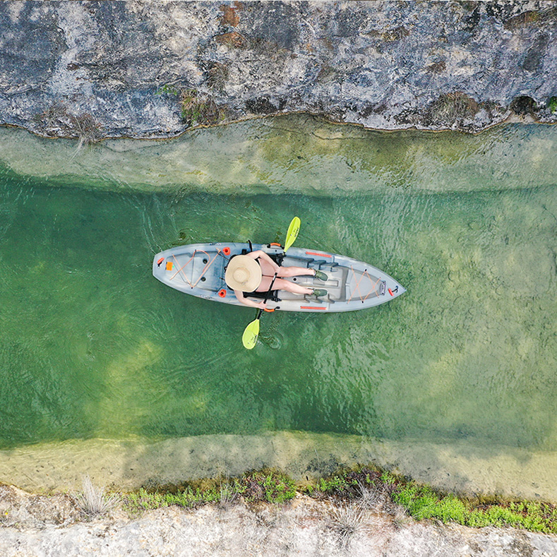 An overhead view of a person kayaking through a narrow, clear green waterway. The kayaker, wearing a wide-brimmed hat, paddles leisurely, with the rocky banks of the water visible on either side. The clear water reveals the natural texture of the riverbed below, creating a tranquil and scenic setting.