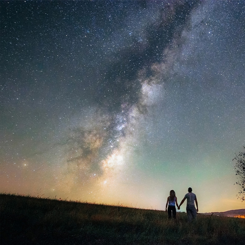 A couple holds hands while standing on a grassy hill, gazing up at the stunning view of the Milky Way stretched across the night sky. The stars and celestial light create a mesmerizing scene, with the vast expanse of the galaxy vividly visible above. The soft glow of distant lights is seen on the horizon, adding a subtle contrast to the natural beauty of the starry sky.