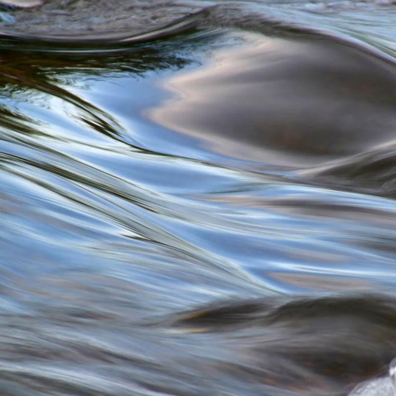 A close-up view of gently flowing water with smooth ripples and soft reflections of the sky and surrounding environment. The water's surface displays shades of blue, grey, and subtle hints of green, creating a calm and serene atmosphere.