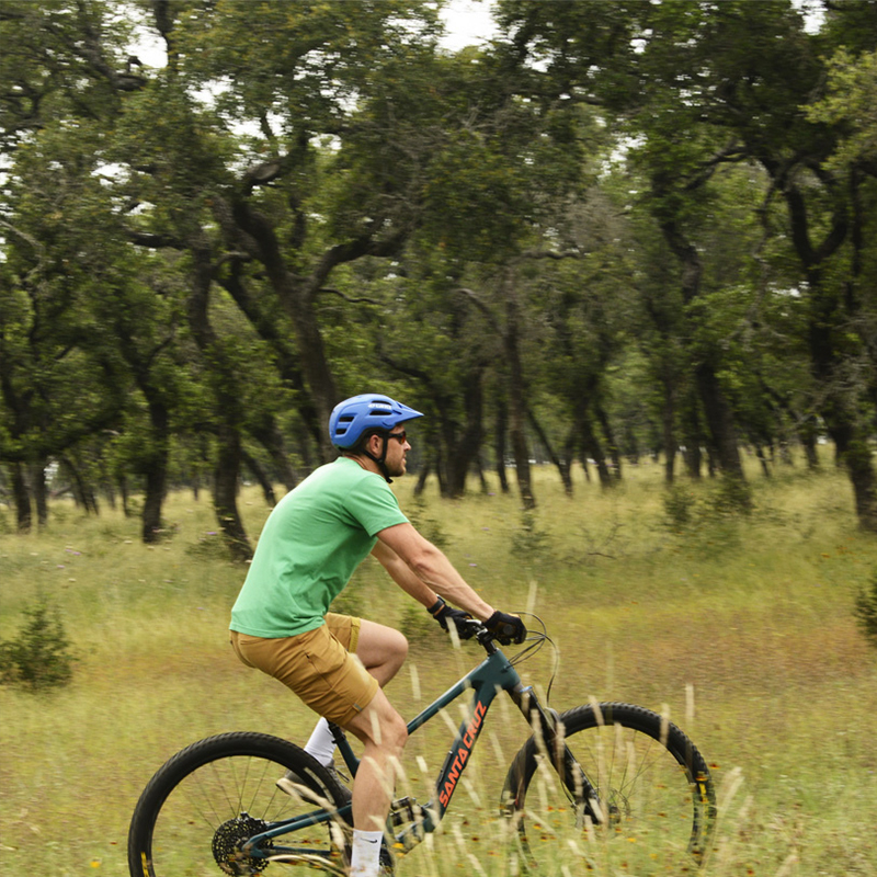 A man wearing a blue helmet rides a mountain bike through a forested area with tall trees and grassy terrain. He is dressed in a green shirt and tan shorts, and the natural setting around him suggests an outdoor adventure in the woods. The trees and greenery provide a calm and shaded environment for his ride.