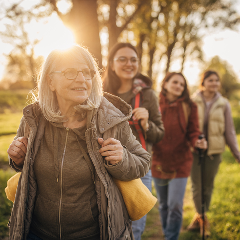 A group of four women hike along a sunlit path, with an older woman in the foreground smiling as she leads the group. Each woman is dressed warmly in jackets, and the soft glow of the setting sun creates a peaceful atmosphere. The background shows trees and greenery, suggesting an outdoor adventure during the golden hour.