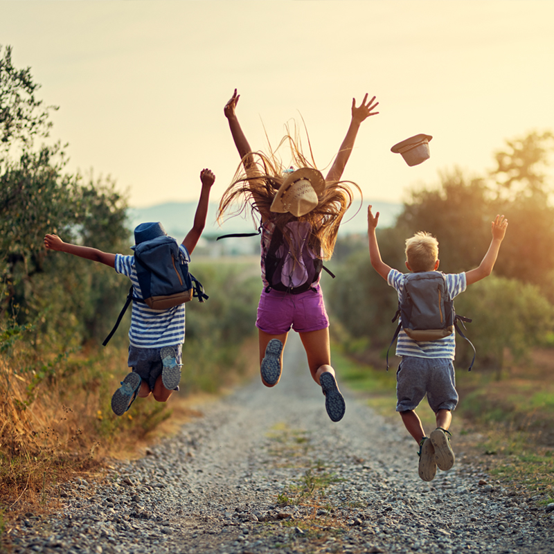 Three children with backpacks jump joyfully in the air on a gravel path, with the sun setting behind them. One child’s hat flies off mid-jump as their long hair waves in the air. The scene is set outdoors, with trees and bushes lining the path, creating a carefree and adventurous atmosphere.