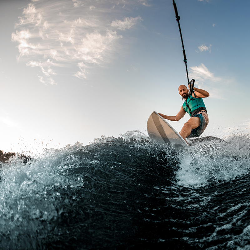 A man wearing a life vest is wakesurfing on a large wave, holding onto a tow rope. The water splashes up around him as he rides the wave, and the sky above is partly cloudy with soft evening light. The dynamic angle of the shot captures the action and energy of the moment.