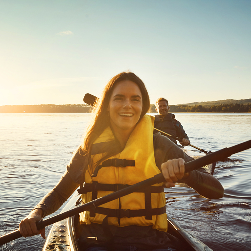 A smiling woman in a yellow life jacket paddles a kayak on a calm lake at sunset, with a man kayaking behind her. The warm sunlight reflects on the water, and the distant shoreline is visible under a clear sky, creating a peaceful and joyful outdoor scene.