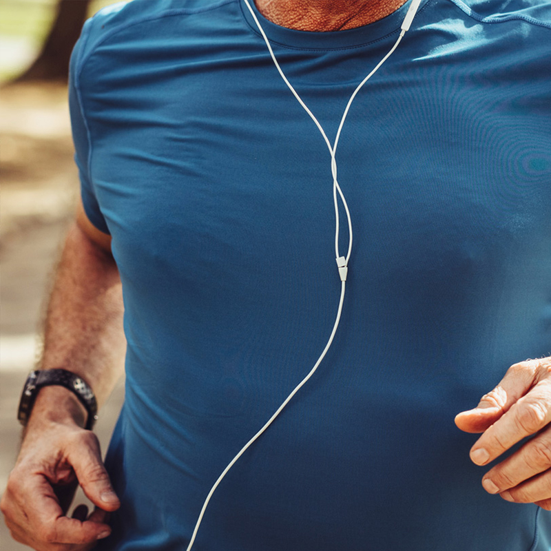 A close-up of a person wearing a blue athletic shirt while jogging. Earphones hang from their neck, suggesting they are listening to music or a podcast. The person’s arm is slightly bent as they run, and a wristwatch is visible on one wrist. The blurred background indicates an outdoor setting, possibly a park or a trail.